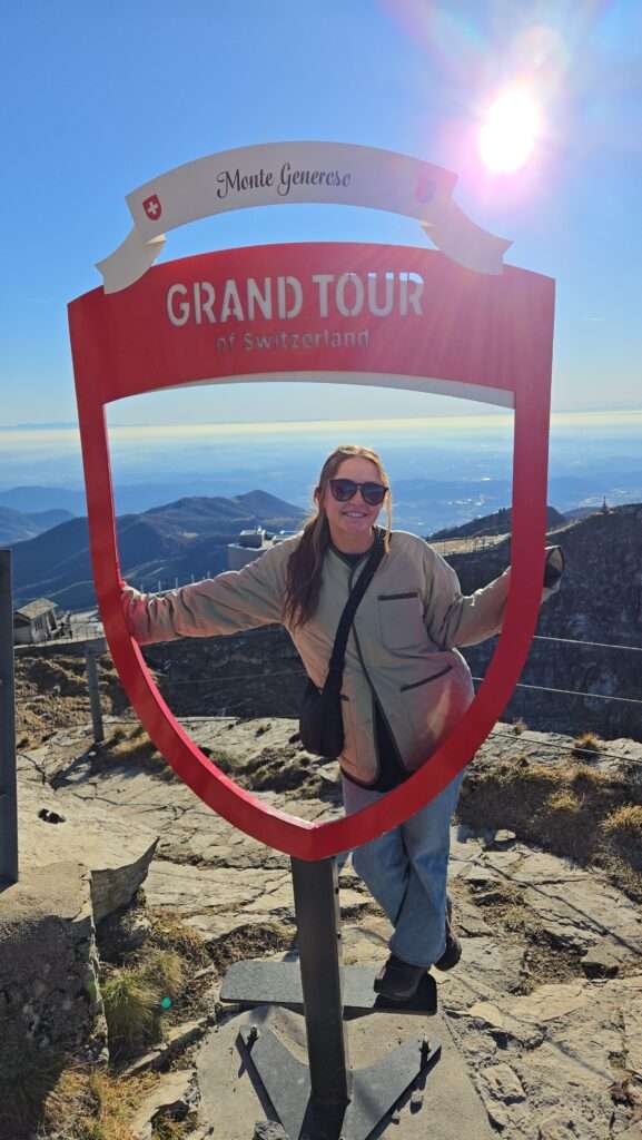 Young woman posing at the Grand Tour of Switzerland sign above Monte Generoso